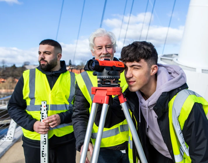 A young student in a high-visibility vest looking through a surveying instrument while the lecturer and another student observe and discuss measurements on a bridge A young student in a high-visibility vest looking through a surveying instrument while the lecturer and another student observe and discuss measurements on a bridge